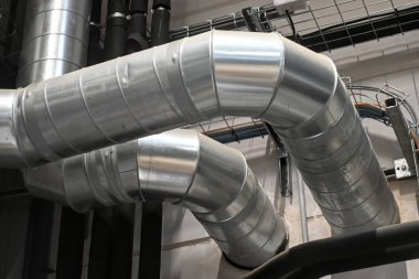 Industrial metal ductwork with bends and joints, part of HVAC system, mounted near ceiling with pipes and cable tray in background.