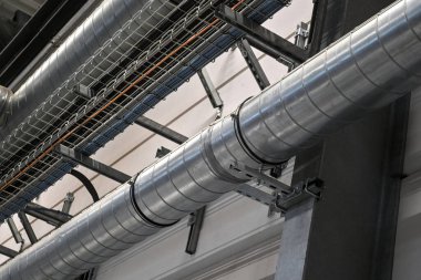 Industrial metal ductwork with bends and joints, part of HVAC system, mounted near ceiling with pipes and cable tray in background.