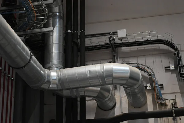 Industrial metal ductwork with bends and joints, part of HVAC system, mounted near ceiling with pipes and cable tray in background.