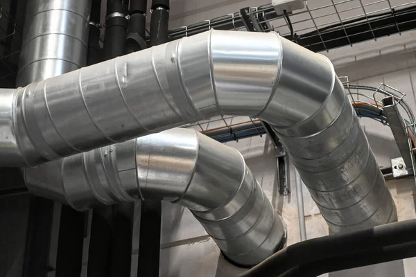 Industrial metal ductwork with bends and joints, part of HVAC system, mounted near ceiling with pipes and cable tray in background.