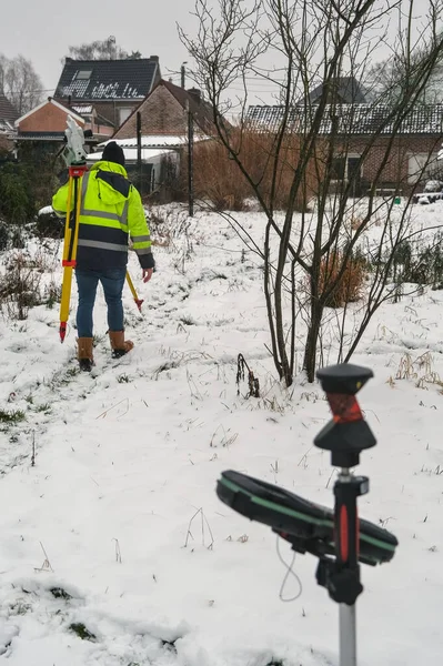 Karlı bir tarlada üç ayaklı bir arazi mühendisi, GNSS alıcısı önplanda, kış koşullarında kadastro sınırını işaretliyor..
