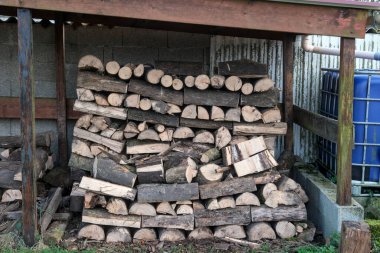 Kırsal bir alanda mavi bir su kabının yanındaki ahşap bir barınağın altına odun yığılmış. Yakacak oduna odaklan..