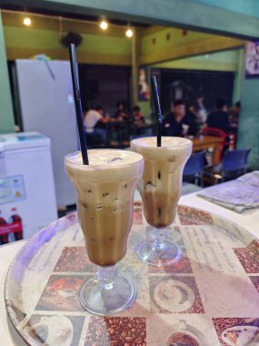 Buzlu kahveyle doldurulmuş, kremalı köpük tabakası ve siyah pipetle kaplanmış iki uzun bardak, desenli bir kafe tepsisinde servis ediliyor..