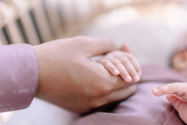 Yetişkin ebeveynler, güvenliği ve derin aile bağlarını vurgulayan yumuşak kumaşa sarılı uyuyan bir bebeğin küçük ve narin parmaklarını sıkıca tutarlar..