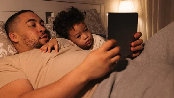Close up view of man and boy lying on bed and reading book on digital tablet. Father and son looking on tablet pc at bedtime.