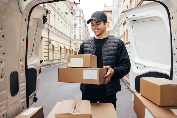 Smiling man courier in uniform standing at car trunk holding boxes preparing for delivery to a customer