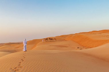 Orta Doğu, Arap Yarımadası, Umman, Ash Sharqiyah North, Bidiya. 25 Ekim 2019. Bir çocuğun Umman çölünde kum tepelerinde deve sallamasını izleyen bir adam..