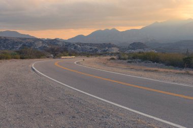 Sıcak, turuncu bir günbatımı gökyüzünün altında kıvrılıp giden boş çöl yolu. 60. yol. Katamarca, Arjantin