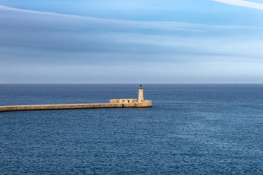Malta 'daki engin mavi Akdeniz ile çevrili taştan bir iskelenin ucunda yeşil tepeli bir deniz feneri barındıran minimalist bir kompozisyon.