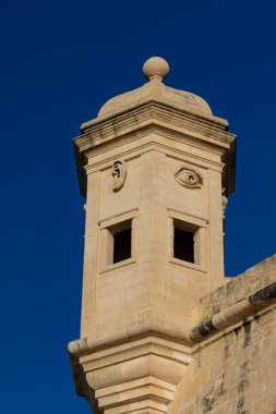Senglea, Malta 'da bir kale duvarındaki' Gardjola 'taş gözetleme kulesinin yüksek açılı, güneşli görüntüsü, koyu mavi gökyüzüne karşı sembolik oyulmuş göz ve kulağı vurguluyor.