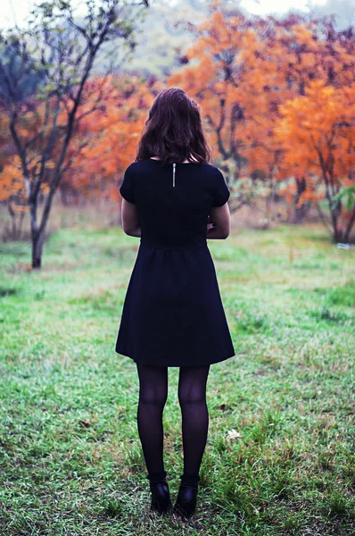 Jovem em um vestido curto preto fica no parque em um backgr — Fotografia de Stock