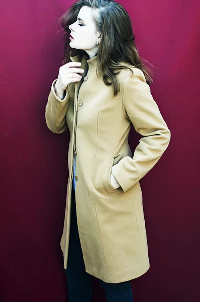 Молодая красивая девушка в молочном пальто на бордовом фоне. Студийная фотография Модная модель в осенней одежде позирует в студии. Портрет модели в позе модного платья — стоковое фото