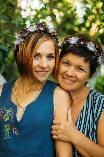Portret uśmiechający się matka i córka są pozowanie w wieńce z kwiatów sztucznych pod gołym niebem na tle drzew. Pojęcie rodziny. Matka i córka związek. — Zdjęcie stockowe