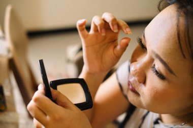 Asyalı bir kadın makyaj pudrası tutarken parmaklarıyla takma kirpik uyguluyor. Kapalı alan arka planının sığ bir derinliğine kırmızı ruj sürüyor..