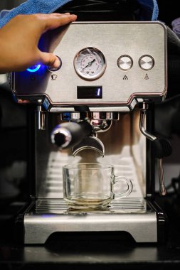 Yakın çekim, bir elin dikey görüntüsü paslanmaz çelik espresso makinesinde parlayan mavi bir düğmeye basarken portafilterden akan sıcak kahve damlalar tepsisindeki cam bardağa akarken.