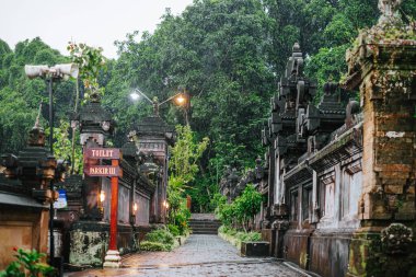 Geleneksel Balili taş tapınak mimarisi Bali, Endonezya 'da asfalt kaplı bir patikayla sınırdır. Aydınlatılmış lambalar ve yemyeşil yapraklar atmosferik, tropikal dış manzarayı tanımlar.