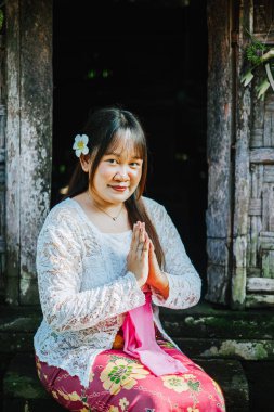 Kebaya 'daki Güneydoğu Asyalı kadın, taşra ahşap bir kapı önünde namaste jesti yapıyor. Bali kültürünü, konukseverliğini ve geleneksel köy hayatını yansıtıyor..