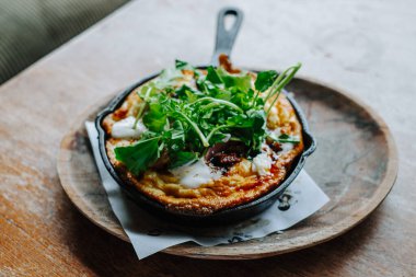 Pişmiş yumurtalı frittata ve taze otlar ahşap bir masada, dökme demir tavada, ev yapımı beslenme, brunch ve otantik mutfak geleneklerini temsil ediyor..