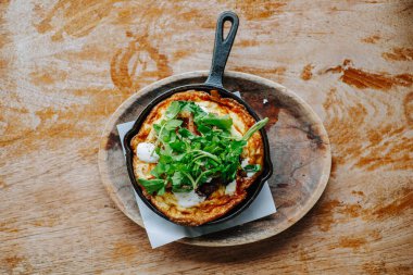 Fırında yumurta frittatasının düz yatışı ve taze otlar dökme demir tavada kırsal ahşap bir masada, ev yapımı beslenme, brunch ve otantik mutfak geleneklerini temsil ediyor.