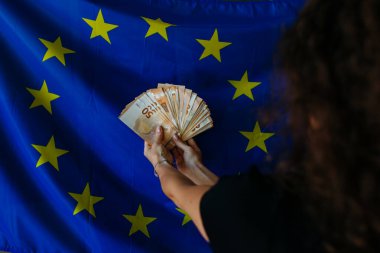 Avrupa Birliği bayrağına karşı avroluk bir deste para tutan kadınların elleri