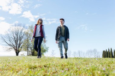 Parkta birlikte yürüyen iki kişinin tam boy görüntüsü.