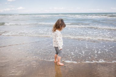 Kumsalda deniz kıyısında duran kahverengi saçlı küçük kızın yan görüntüsü.