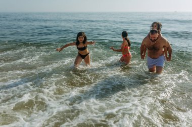 Güneşli bir günde dalgalarla oynayan baba ve üç kızın yüksek açılı görüntüsü.