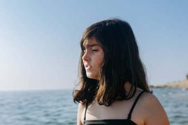 Parlak gün ışığında gözlerini kısarak bakan genç bir kızın yakın plan portresi. Sahilde duruyor.