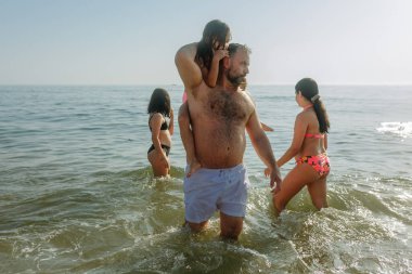 Baba ve üç kızı sahilde dalgalarla oynuyorlar. Küçük çocuk bir ebeveynin boynunda oturuyor.