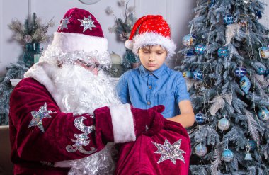 Noel Baba ve kırmızı şapkalı bir çocuk Noel ağacının yanında oturuyorlar, çocuk Noel Baba 'nın çantasına bakıyor ve hediyelerini çıkarıyor, Noel ve yeni yıl, kış tatilleri..