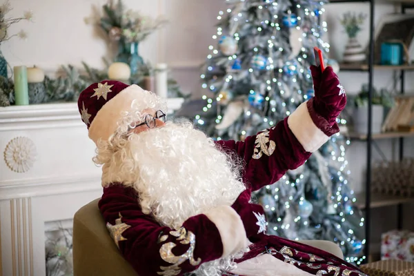 Santa Claus in a red hat and a big curly white beard, says Christmas greetings on his smartphone, he is wearing glasses by the New Year tree, a portrait of a New Year character, an animator in a Santa