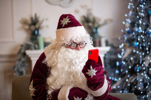 Santa Claus in a red hat and a big curly white beard, says Christmas greetings on his smartphone, he is wearing glasses by the New Year tree, a portrait of a New Year character, an animator in a Santa