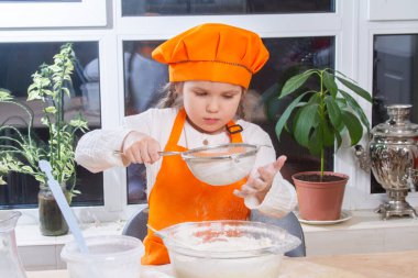 A little cute girl in an orange chef costume breaks a raw egg and pours it into a bowl of flour, a child prepares and kneads dough for baking, daughter helps to cook in the kitchen.