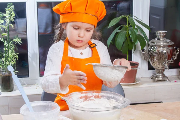 A little cute girl in an orange chef costume breaks a raw egg and pours it into a bowl of flour, a child prepares and kneads dough for baking, daughter helps to cook in the kitchen.