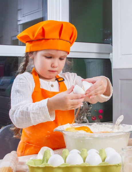 A little cute girl in an orange chef costume breaks a raw egg and pours it into a bowl of flour, a child prepares and kneads dough for baking, daughter helps to cook in the kitchen.