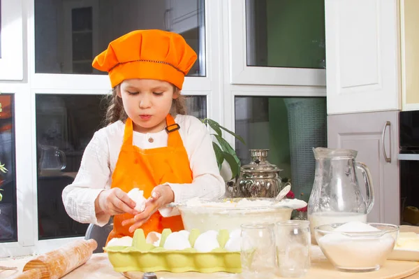 A little cute girl in an orange chef costume breaks a raw egg and pours it into a bowl of flour, a child prepares and kneads dough for baking, daughter helps to cook in the kitchen.