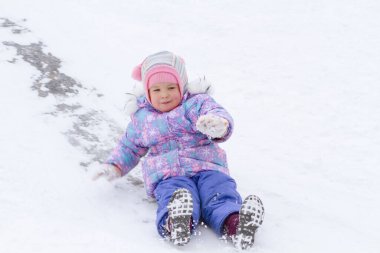 Mor tulumlu küçük bir kız buz kaydırağından kayıyor, kış çocuk oyunları, kışın parkta bir çocuk..