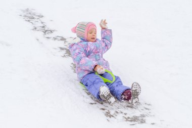 Mor tulumlu küçük bir kız buz kaydırağından kayıyor, kış çocuk oyunları, kışın parkta bir çocuk..