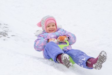 Mor tulumlu küçük bir kız buz kaydırağından kayıyor, kış çocuk oyunları, kışın parkta bir çocuk..