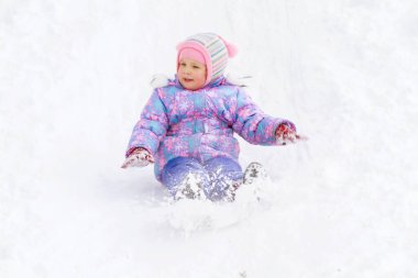 Mor tulumlu küçük bir kız buz kaydırağından kayıyor, kış çocuk oyunları, kışın parkta bir çocuk..
