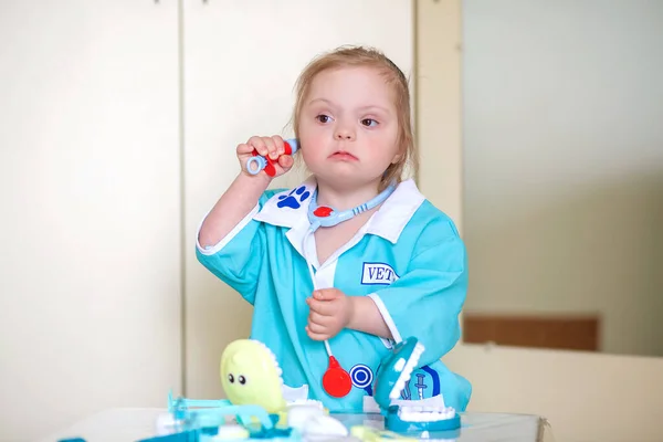 Down syndrome in a little girl, a child in a doctor\'s costume playing with a doll and with medical instruments, disabled children, genetic mental illness, an extra chromosome in humans.