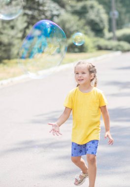Mutlu bir kız büyük sabun köpükleri yakalar, bir çocuk sokakta oynar, havada hafif balonlar süzülür..