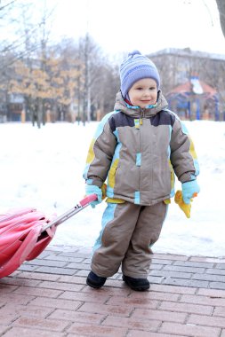 Renkli bir kış kıyafeti giymiş genç bir çocuk, karlı bir parkı keşfediyor, patika boyunca canlı pembe bir şemsiye çekiyor, buzlu ağaçlar ve kış mucizeleriyle çevrili..
