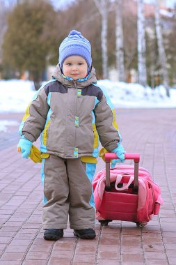Renkli bir kış kıyafeti giymiş neşeli genç bir çocuk, karla kaplı yeşil bir yol boyunca pembe bir bavulu sürüklüyor ve macera ruhunu yakalıyor..