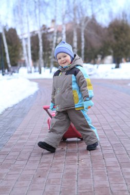 Renkli bir kar elbisesi giyen neşeli bir çocuk, kış manzarasıyla çevrili bir kaldırım taşı patikasında parlak kırmızı bir kızak çekiyor. Çocuk, onlar karlı havanın tadını çıkarırken gülümsüyor..