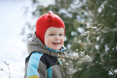 Neşeli bir çocuk açık havayı sever. Renkli bir ceket ve kırmızı bir şapka giyer. Etrafı karlı zemin ve parlak güneş ışığı altında yemyeşil yapraklarla çevrili, etrafı keşfederken gülümsüyor..
