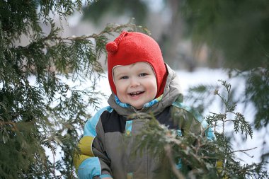 Parlak kırmızı şapkalı neşeli genç bir çocuk karla kaplı Evergreen 'lerin arasında oynarken gülümsüyor, güneşli bir günde kışın neşeli ruhunun tadını çıkarıyor..