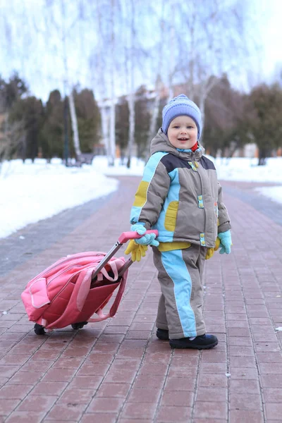 Renkli kış giysileri içinde gülümseyen genç bir çocuk donmuş bir yolda yürüyor, arkasında neşeli pembe bir bavul taşıyor. Karlı ağaçlar büyülü bir kış manzarası yaratır..