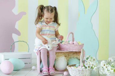 Çizgili elbiseli neşeli çocuk paskalya pastel Paskalya yumurtaları ve bahar neşesini vurgulayan şenlikli süslemelerle çevrili neşeli bir Paskalya sahnesinde beyaz bir tavşanla etkileşime girer..