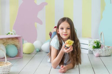 Büyüleyici sahne, yumuşak bir civcivi kucağında tutan, açık tahta zeminde yatan genç bir kızı anlatıyor. Sıcak bir şekilde gülümsüyor. Etrafı pastel süslemelerle çevrili. Neşeli bir bahar havası katıyor..